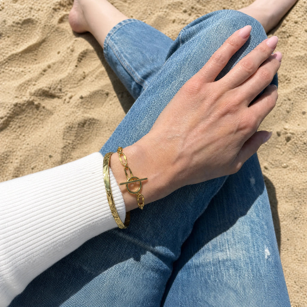 The perfect bracelet duo. The So Hum Gold Cuff Bracelet worn with the Burning Desire Gold OT Fastening Chain Bracelet. By Frequency Paris Ibiza. Worn with a slimline white sweater and blue jeans by the beach.