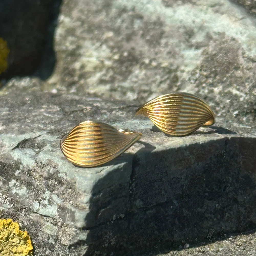 Frequency Paris-Ibiza Eternal Wave Gold Hoop Earrings placed on organic rock by the beach, highlighting flowing wave design and everyday elegance