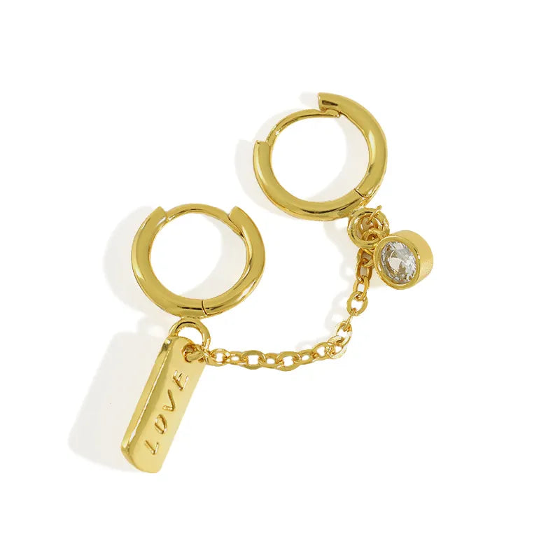 Close-up of Frequency Paris-Ibiza Self Love Huggie Chain Earrings in 18K gold-plated sterling silver. Double huggie design with chain, sparkling zircon crystal on one huggie and ‘love’ tag on the other.