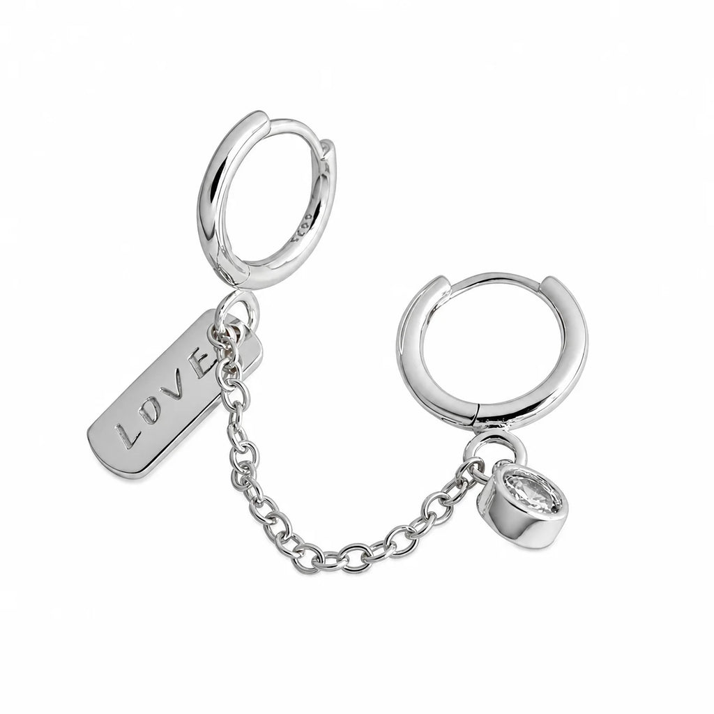 Frequency Paris-Ibiza Self Love Huggie Chain Earrings in rhodium-plated sterling silver on white background. Double huggie with chain, crystal drop gemstone, and ‘love’ tag. Perfect for day-to-night wear.