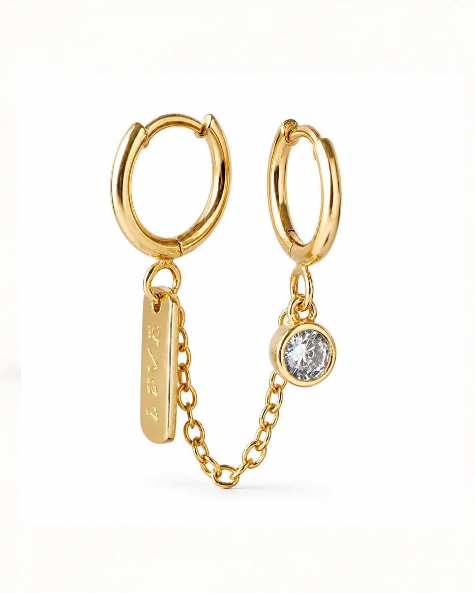 Close-up of Frequency Paris-Ibiza Self Love Huggie Chain Earrings in 18K gold-plated sterling silver. Double huggie design with chain, sparkling zircon crystal on one huggie and ‘love’ tag on the other